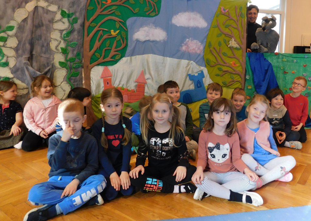 Grupa dzieci siedzą na podłodze , w tle dekoracja teatralna